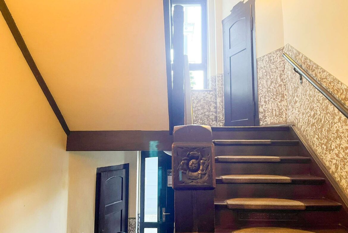 Interior staircase with dark wood steps, ornate carved newel post, floral wallpaper on lower wall, and a door at the top with natural light streaming in