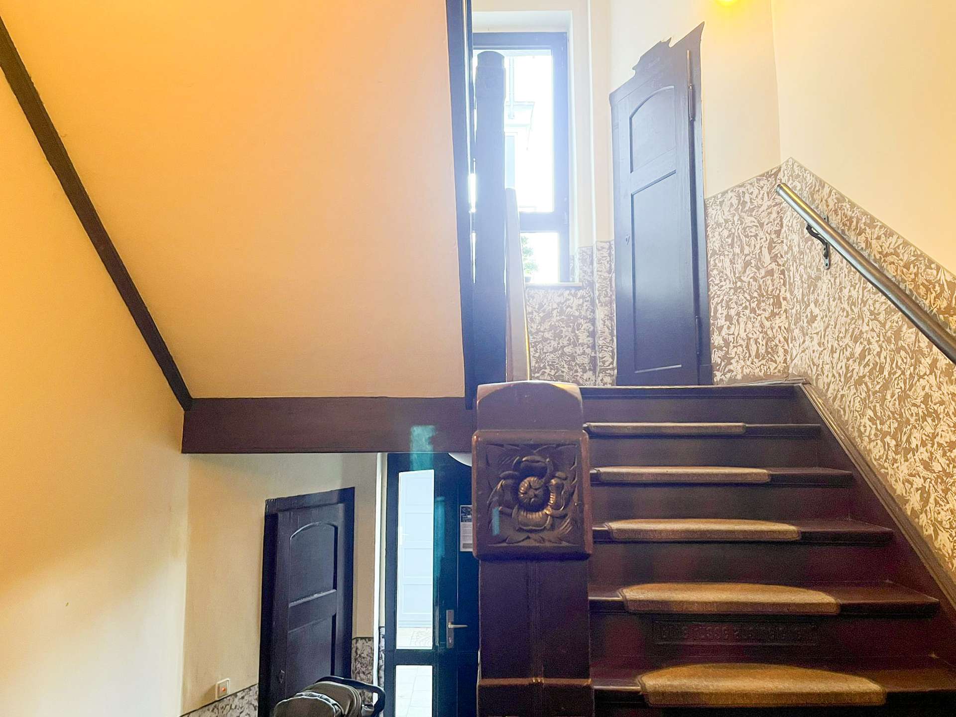 Interior staircase with dark wood steps, ornate carved newel post, floral wallpaper on lower wall, and a door at the top with natural light streaming in