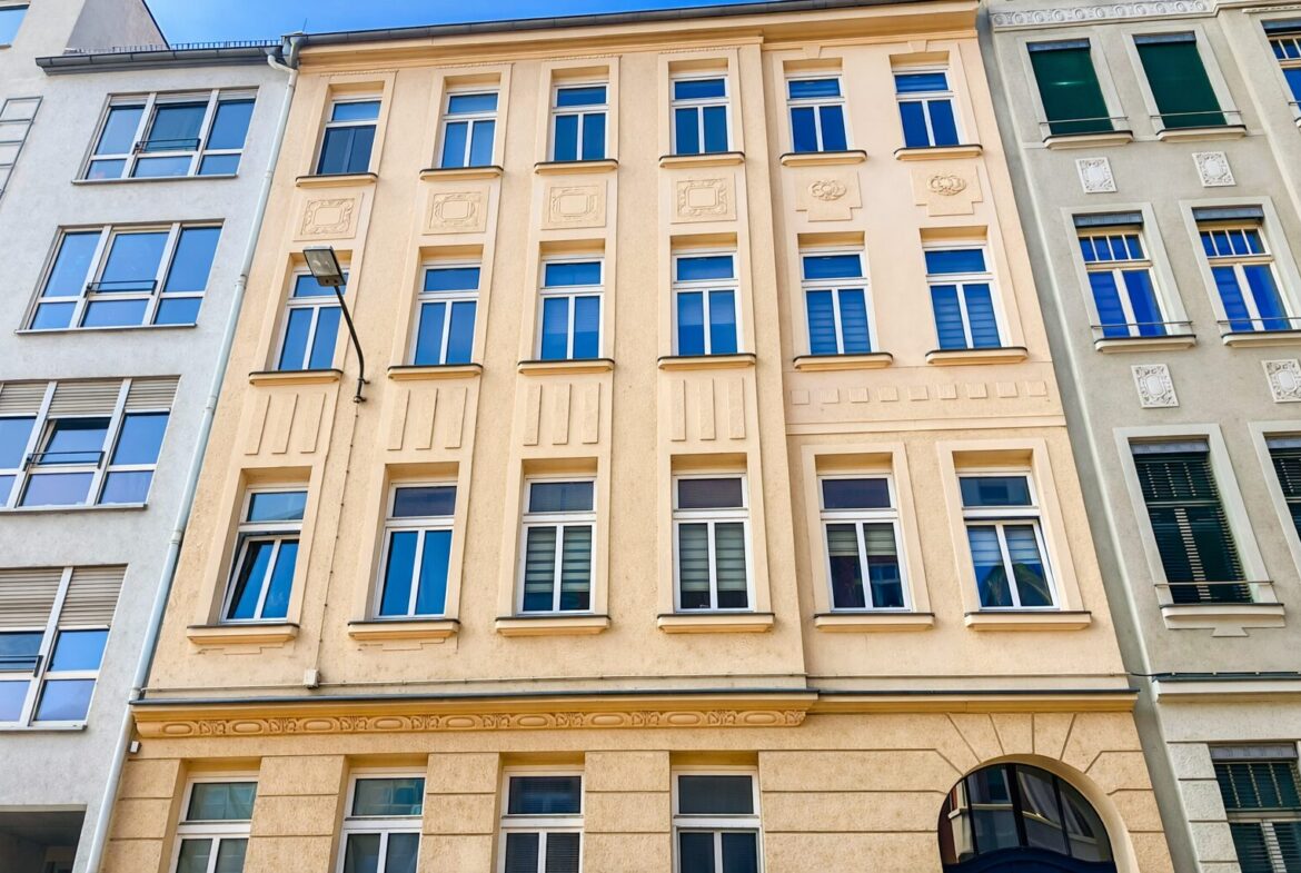Beige, classical building facade with symmetrical windows and decorative panels, flanked by gray buildings; a street lamp extends from the left.