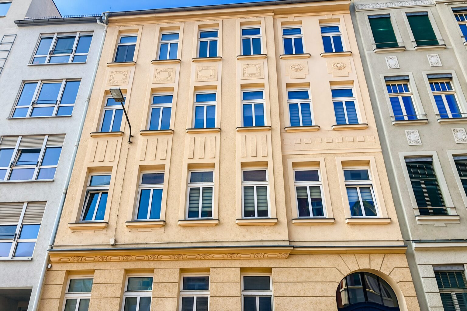 Beige, classical building facade with symmetrical windows and decorative panels, flanked by gray buildings; a street lamp extends from the left.