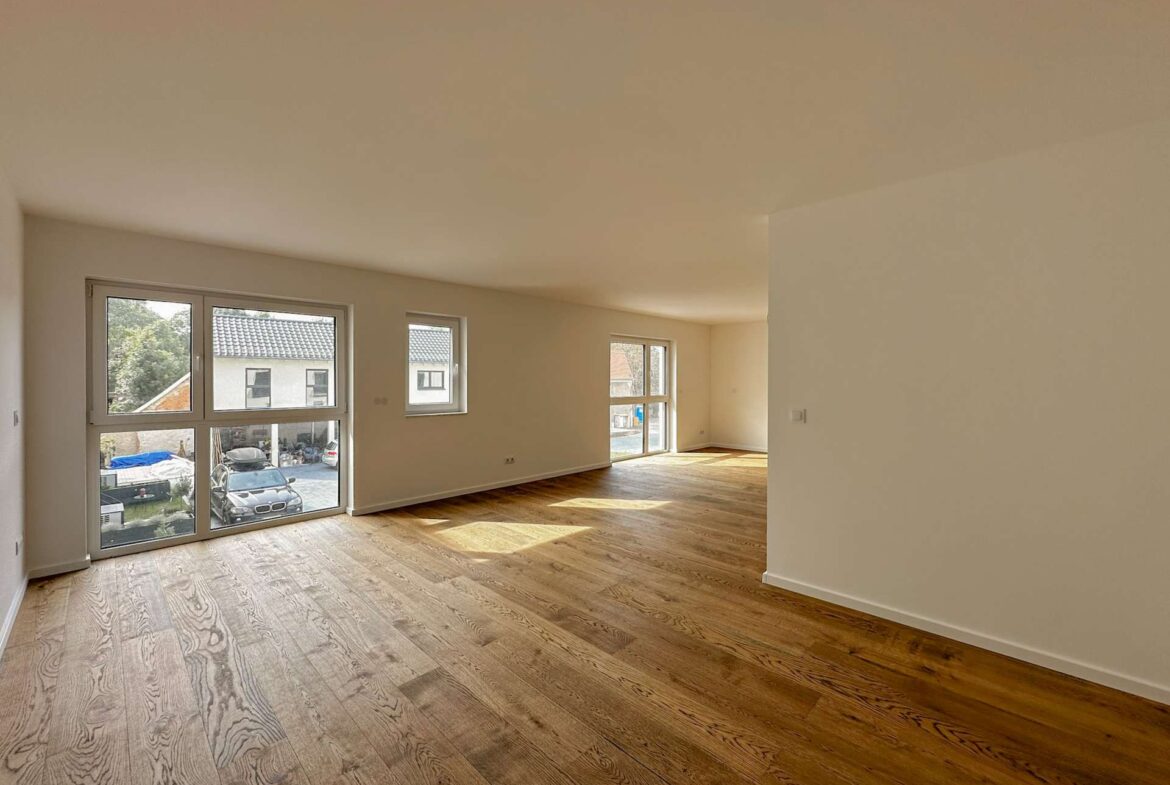 Open, empty living room with wood floors and large windows letting in natural light, neutral walls, and a curved partition.