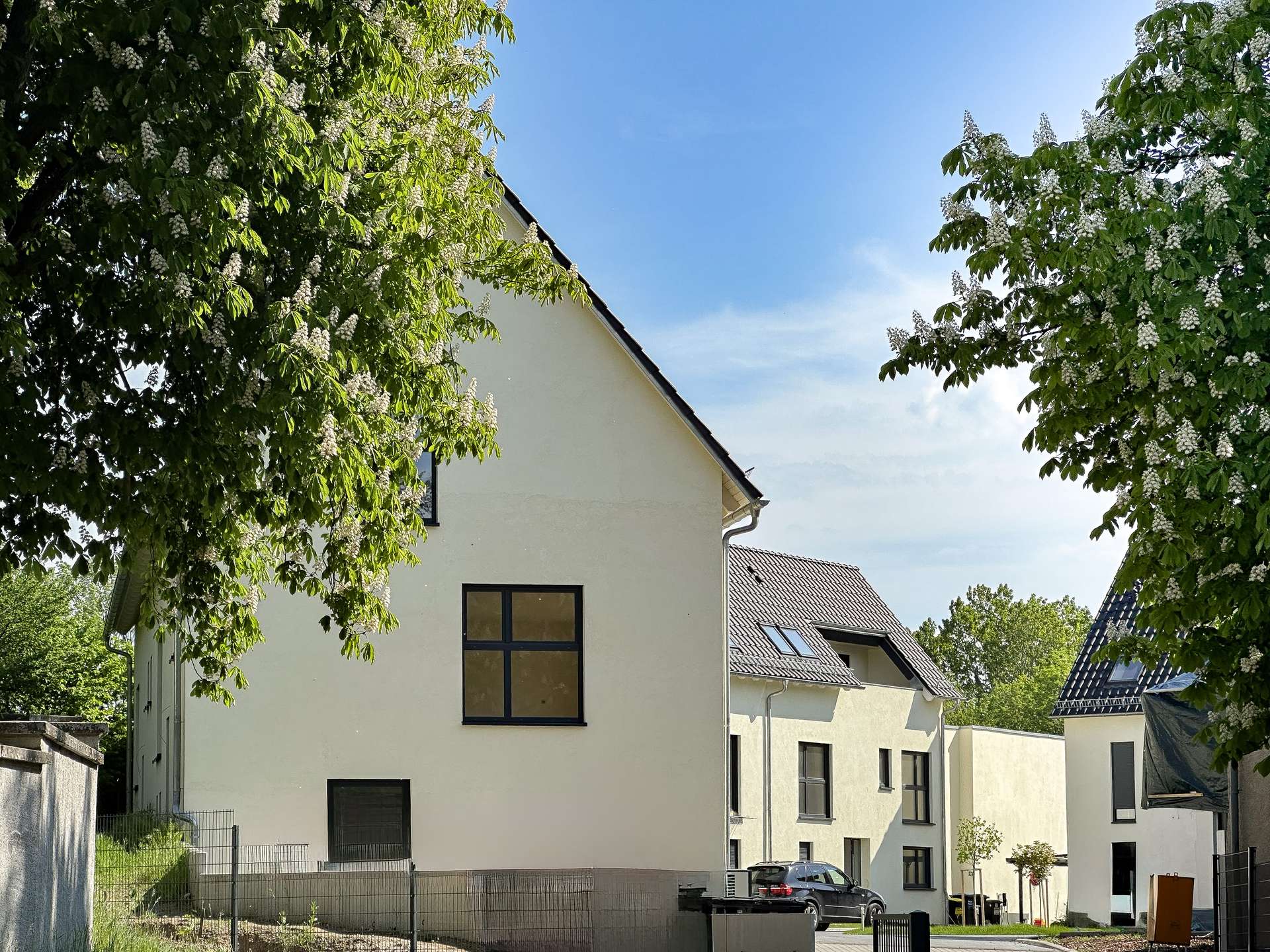 Modern white townhouse complex with black-framed windows, under a blue sky, cars in the driveway and flowering trees nearby.