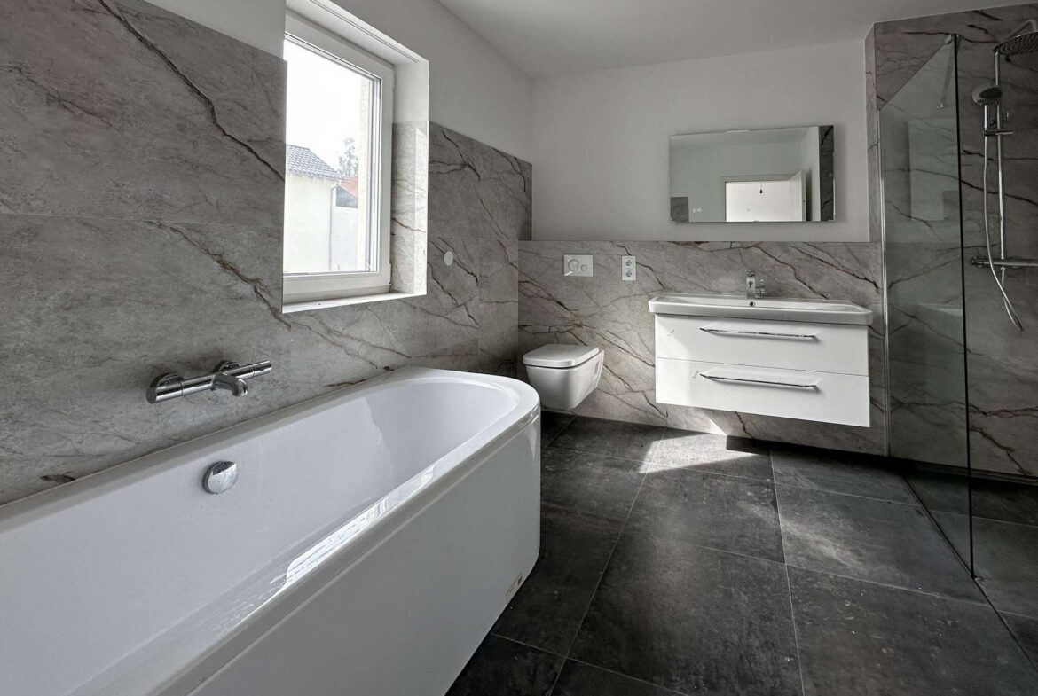 Modern bathroom with gray marble walls and floors, white freestanding tub, wall-mounted vanity, and glass shower on the right.