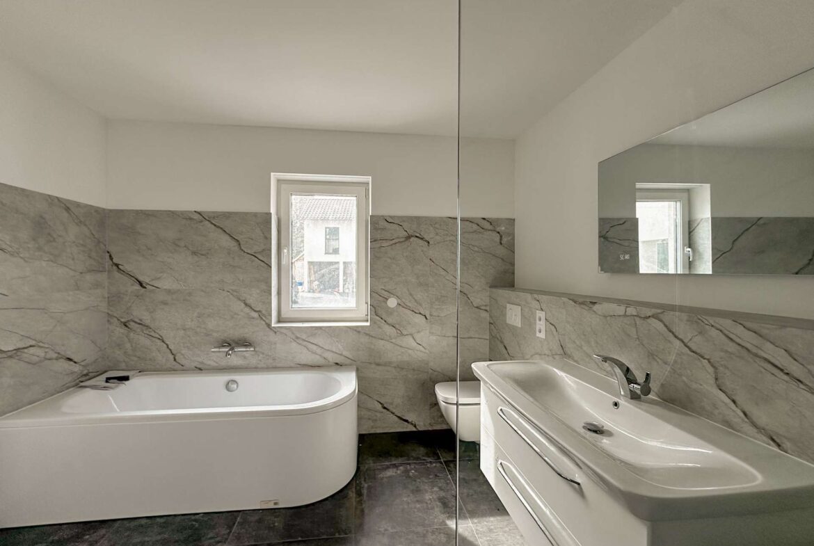 Modern bathroom with white tub on left, marble tiled walls, small window, and a white sink with chrome faucet on the right, mirrored by a large wall mirror.