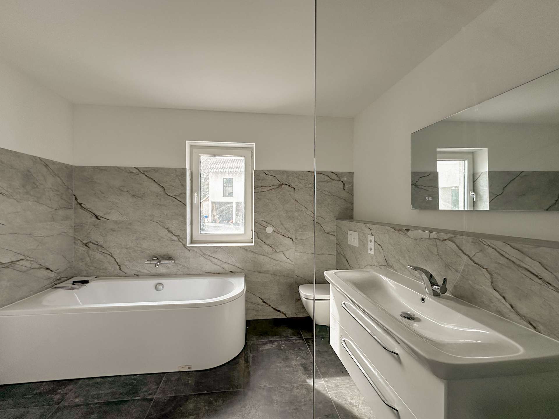 Modern bathroom with white tub on left, marble tiled walls, small window, and a white sink with chrome faucet on the right, mirrored by a large wall mirror.