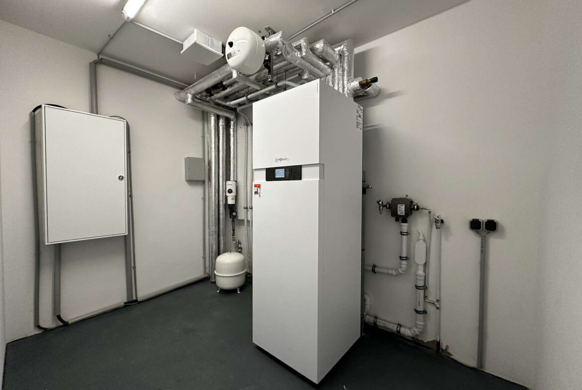 Industrial boiler room with white vertical boiler, insulated pipes, and an expansion tank; utility controls on the unit present.