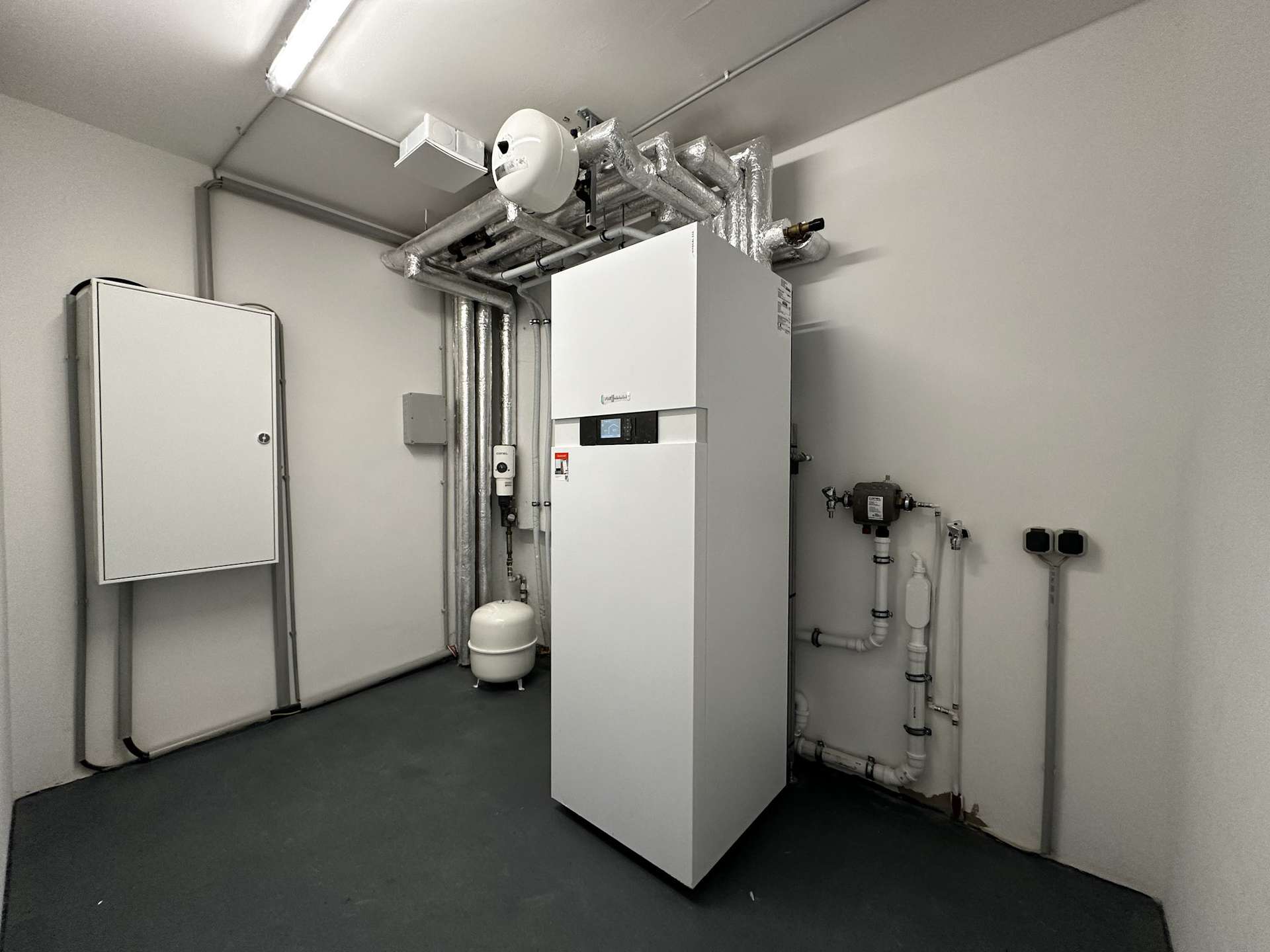 Industrial boiler room with white vertical boiler, insulated pipes, and an expansion tank; utility controls on the unit present.