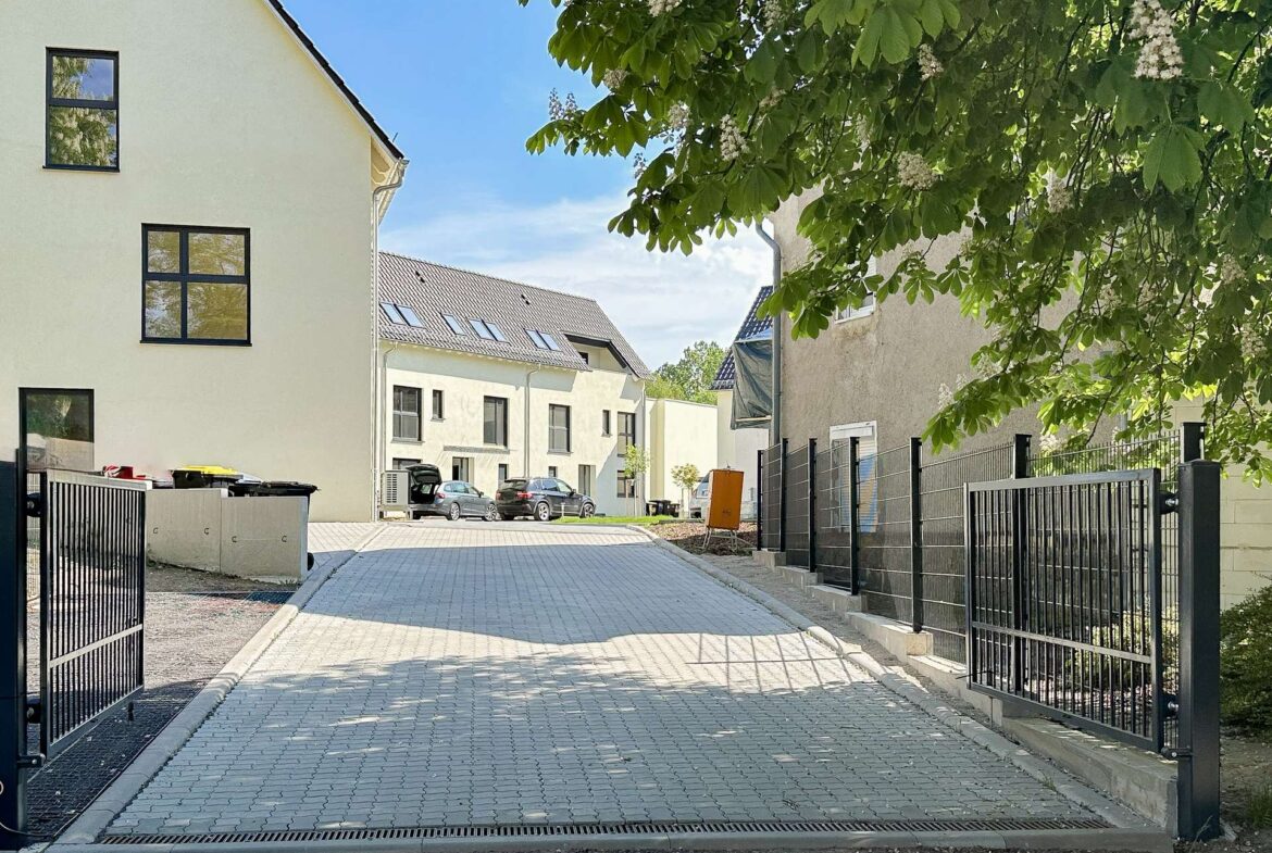 Residential street with modern white houses, parked cars, and a large leafy tree with white blossoms overhead kaped by fences on both sides.