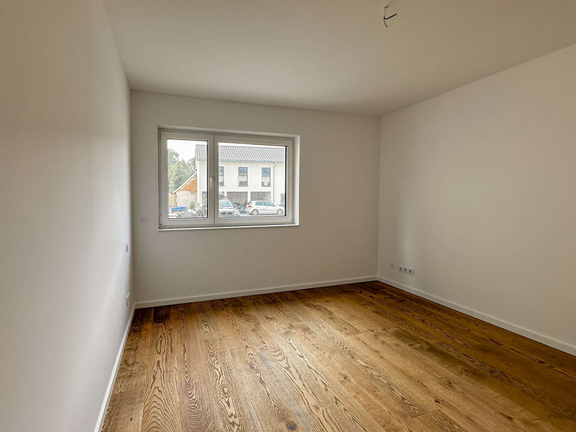 Empty white room with a window letting in light and oak hardwood flooring.