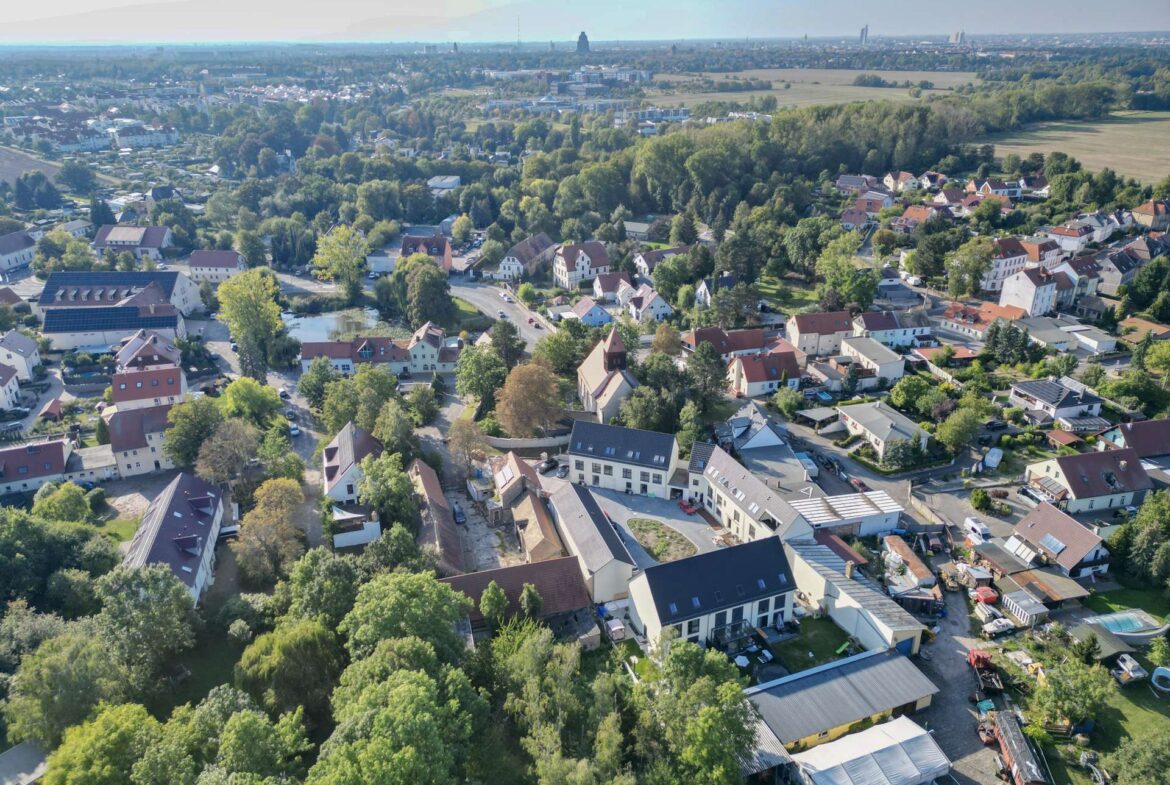 Aerial view of a suburban town with red-roof houses, tree-lined streets, and fields in the distance.