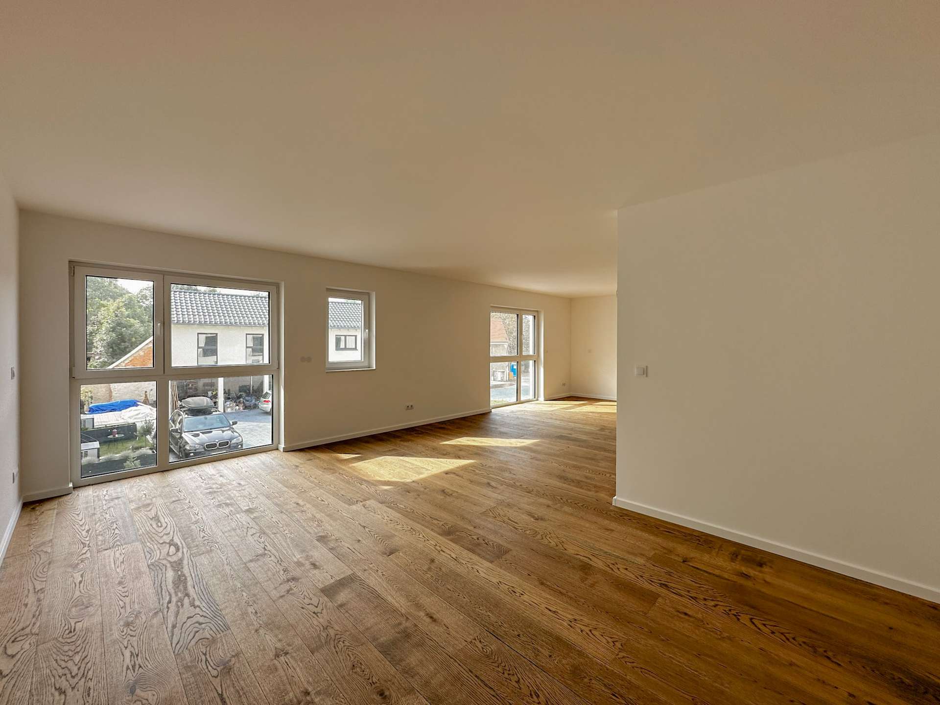 Bright, empty living room with wide glass doors and windows, light wood flooring and neutral walls viewed from inside the space.