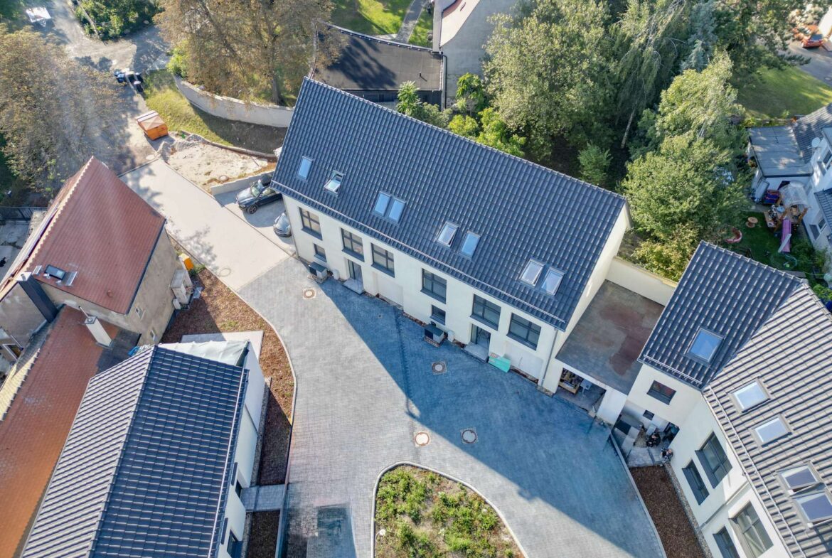 Aerial view of a modern residential complex with white buildings, grey tiled roofs, a central paved courtyard, and surrounding green trees.