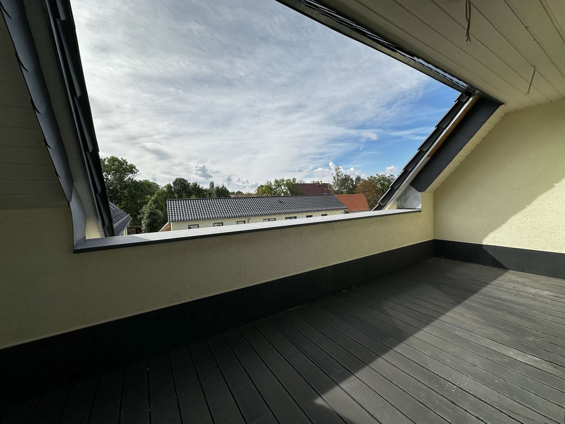 Open balcony with dark wooden decking and pale beige walls, viewing a cloudy sky and distant rooftops through a large triangular opening.