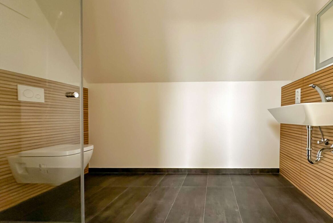 Modern bathroom with a wall-mounted toilet, wood-grain panels, glass shower door, and white sink under warm lighting.