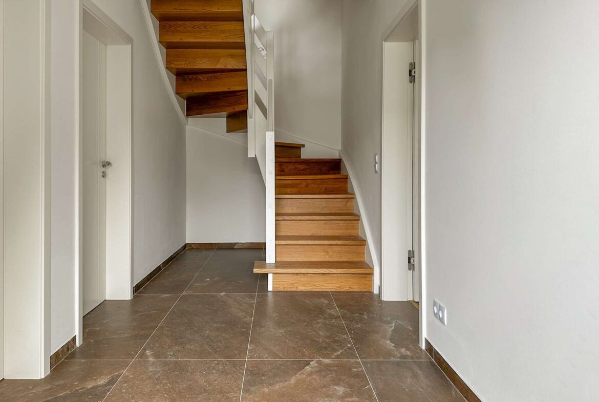 Narrow hallway with a wooden staircase winding up from the center, white walls, and doors along the sides.