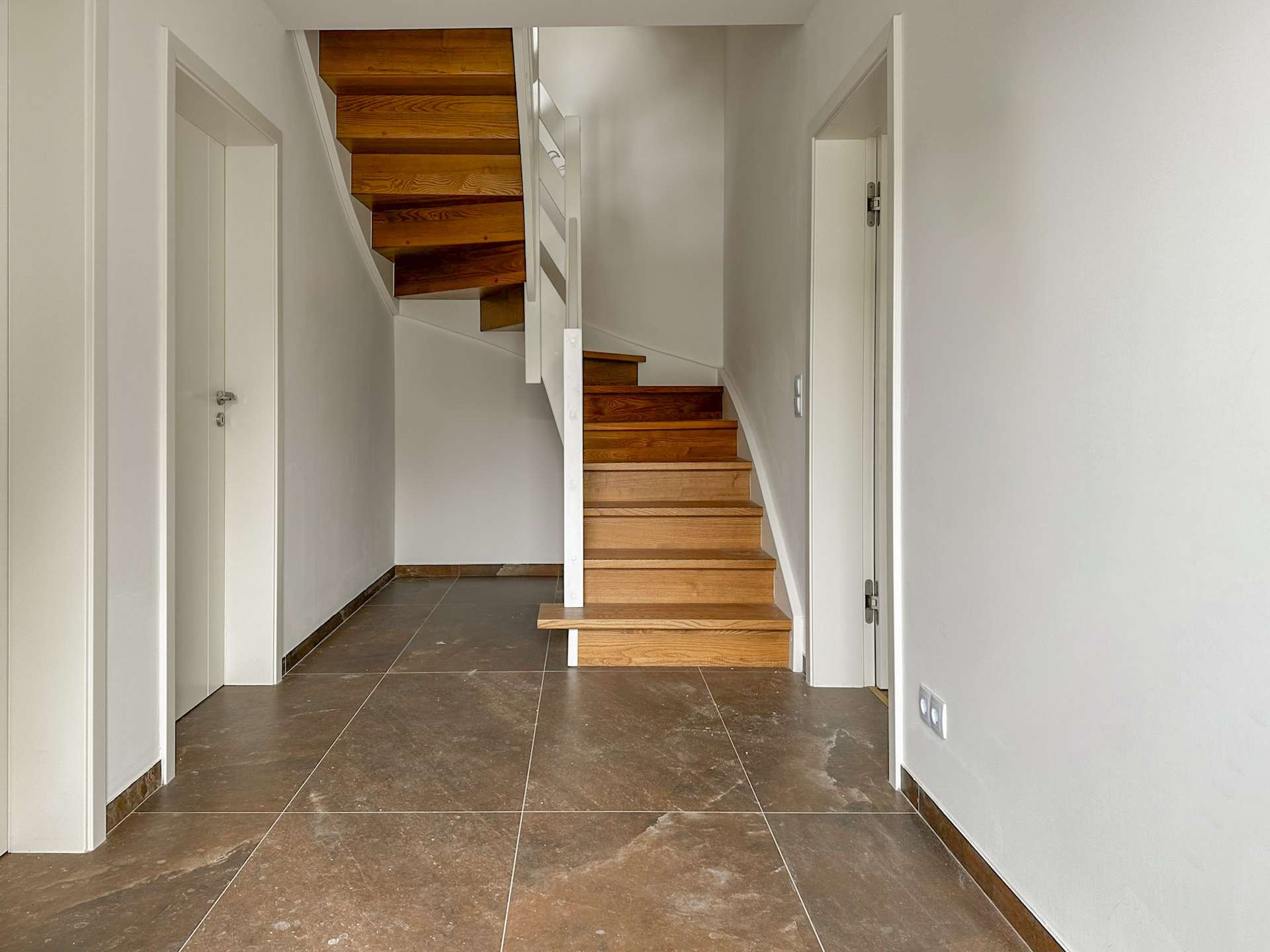 Narrow hallway with a wooden staircase winding up from the center, white walls, and doors along the sides.