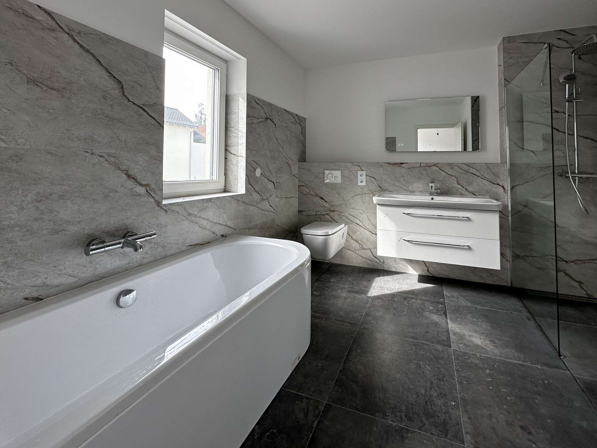 Modern bathroom with gray marble walls and floor, built-in tub left, toilet, white vanity, mirror, and glass-enclosed shower.