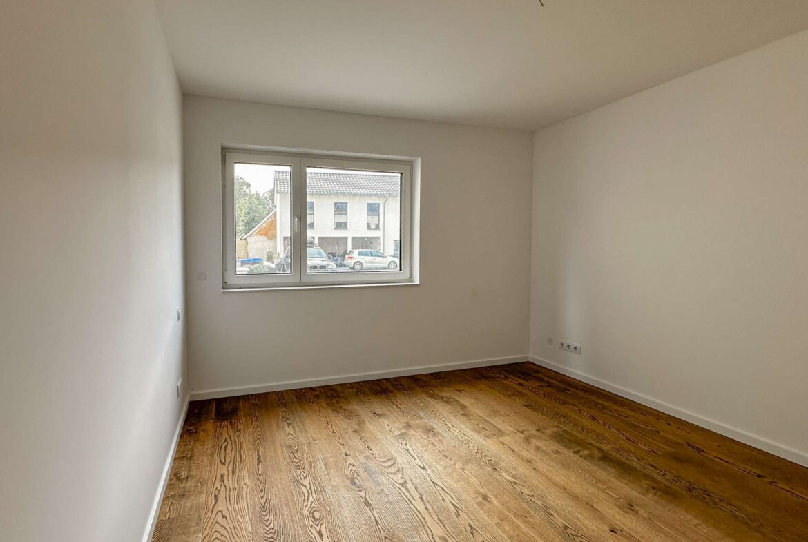 Empty, unfurnished room with white walls, a wooden floor, and a window looking out at neighboring houses and parked cars.