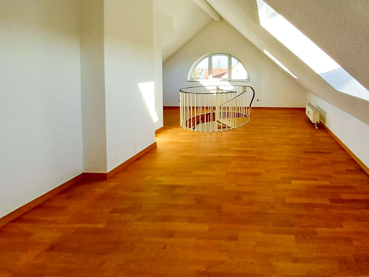 Bright attic interior with warm wooden floor, circular staircase railing at center, arched window, and radiator on the right side.