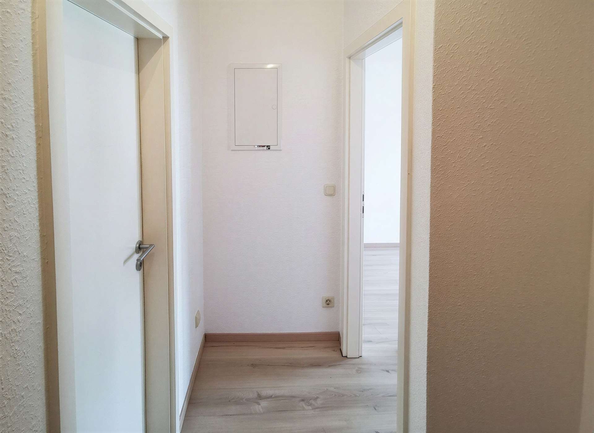 Narrow hallway with light wood flooring, white doors and trim, textured walls, and an open doorway at the far end showing a bright room.