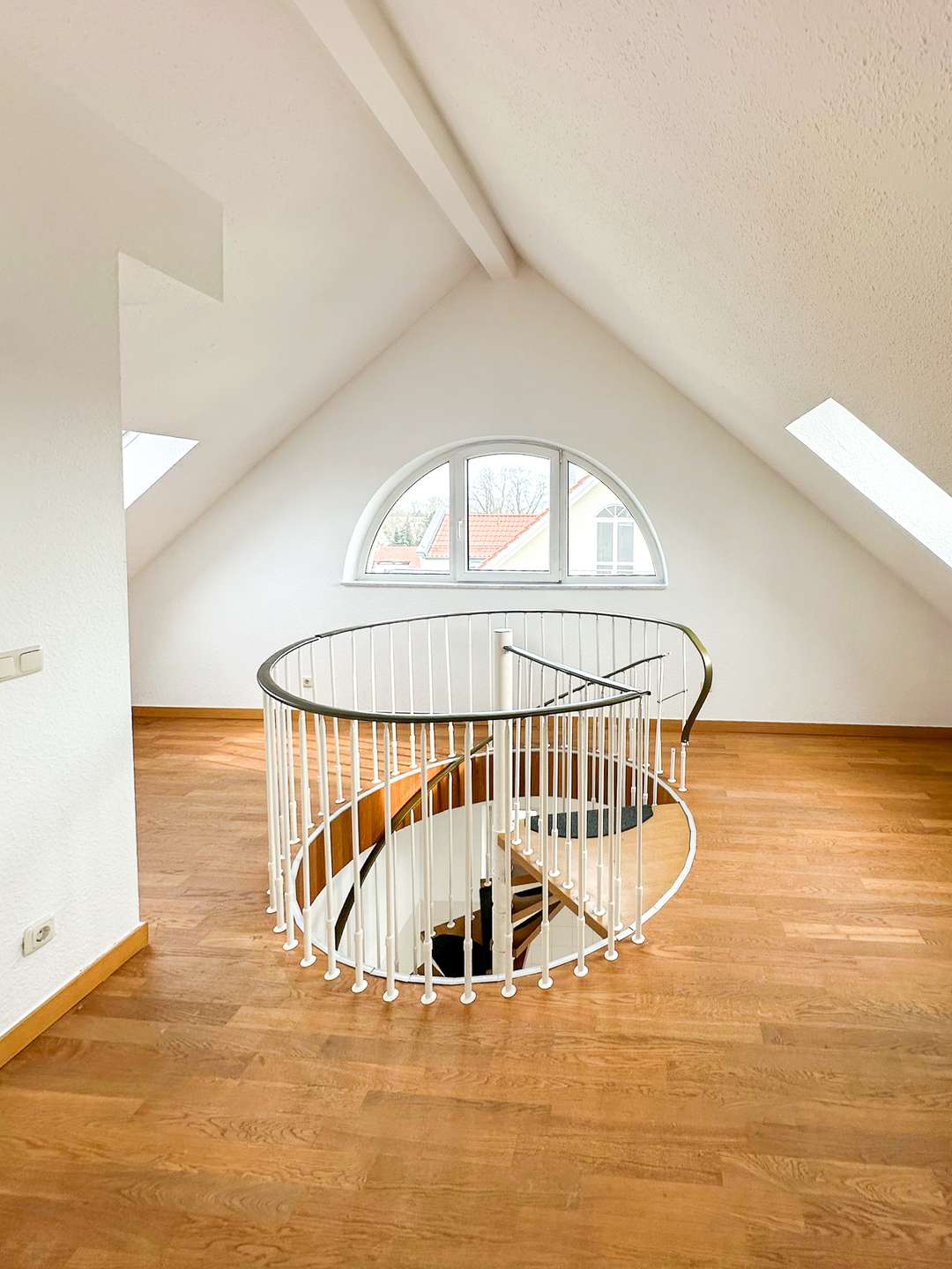 Spiral staircase in a bright attic with white railing, wooden floor, and a semicircular window at the end of the hall.