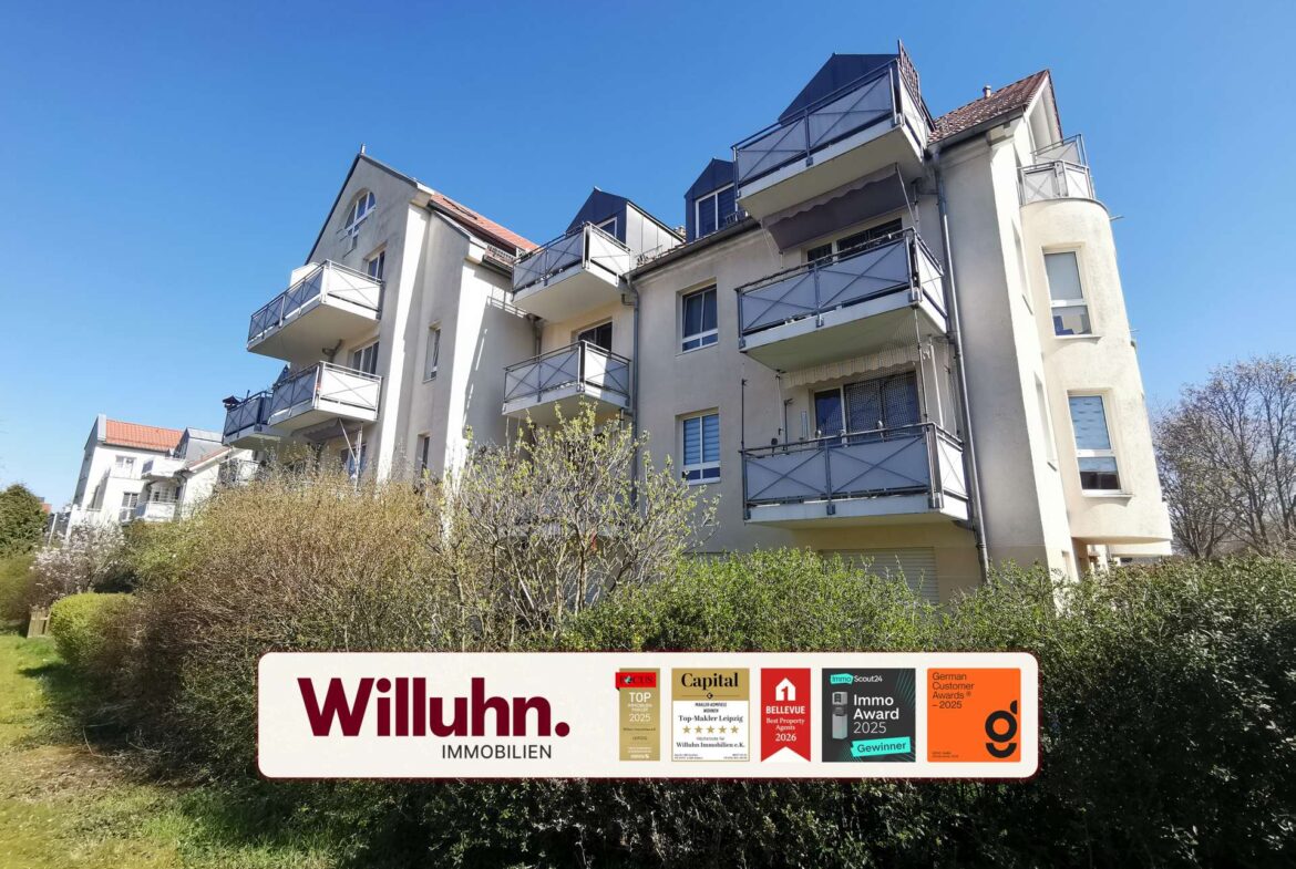 Apartment building with many balconies facing a clear blue sky; a large Willuhn Immobilien real estate sign with award stickers in the foreground.