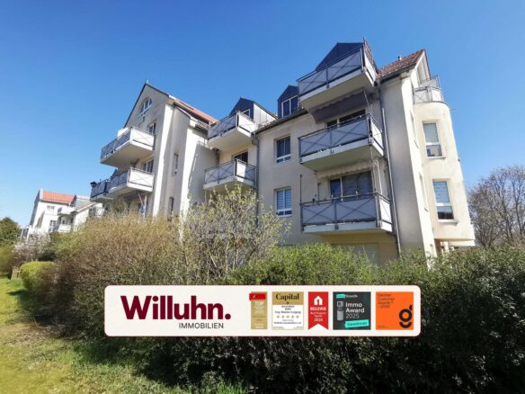 Apartment building with many balconies facing a clear blue sky; a large Willuhn Immobilien real estate sign with award stickers in the foreground.