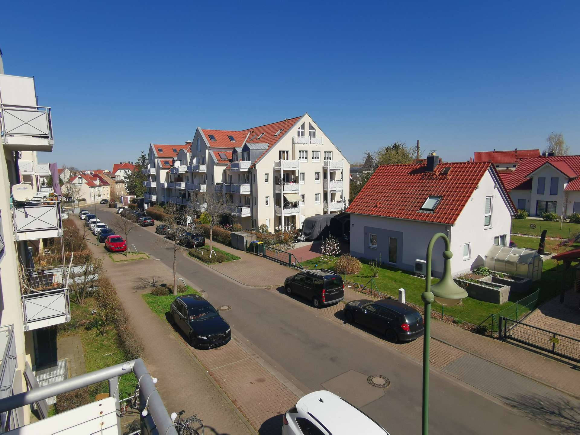Residential street with white apartment buildings and red-tiled roofs under a bright blue sky; parked cars line the curbside.