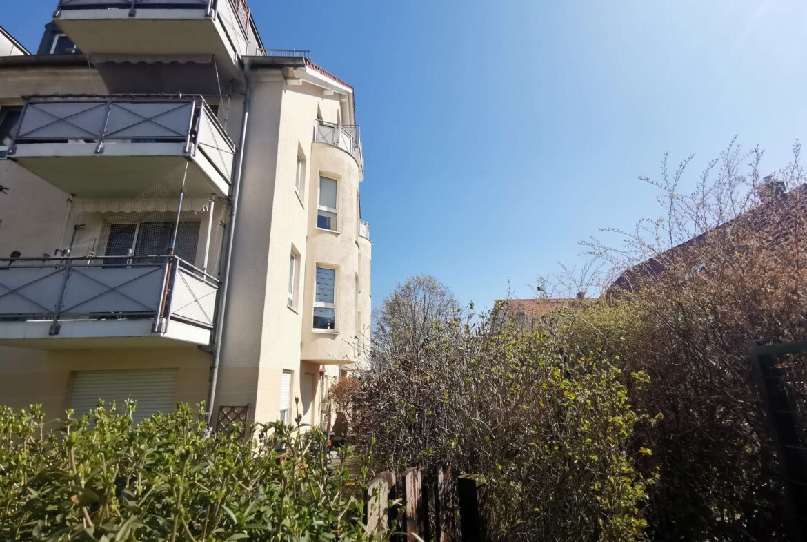 Apartment building with multiple balconies along a beige facade, under a bright blue sky, with a leafy hedge in the foreground.