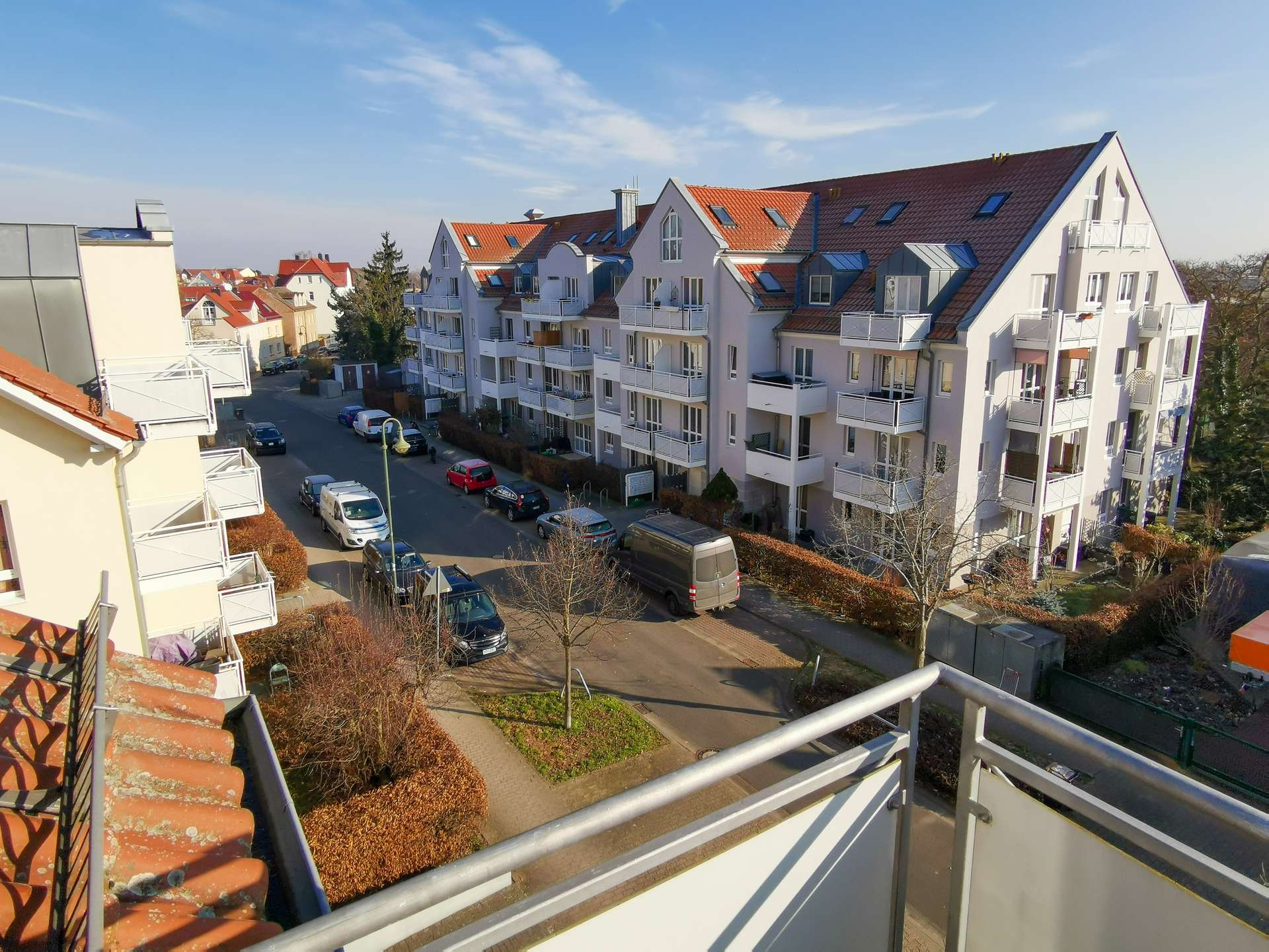 Balcony view over modern white apartment buildings with red-tiled roofs, parked cars, and a sunny blue sky.