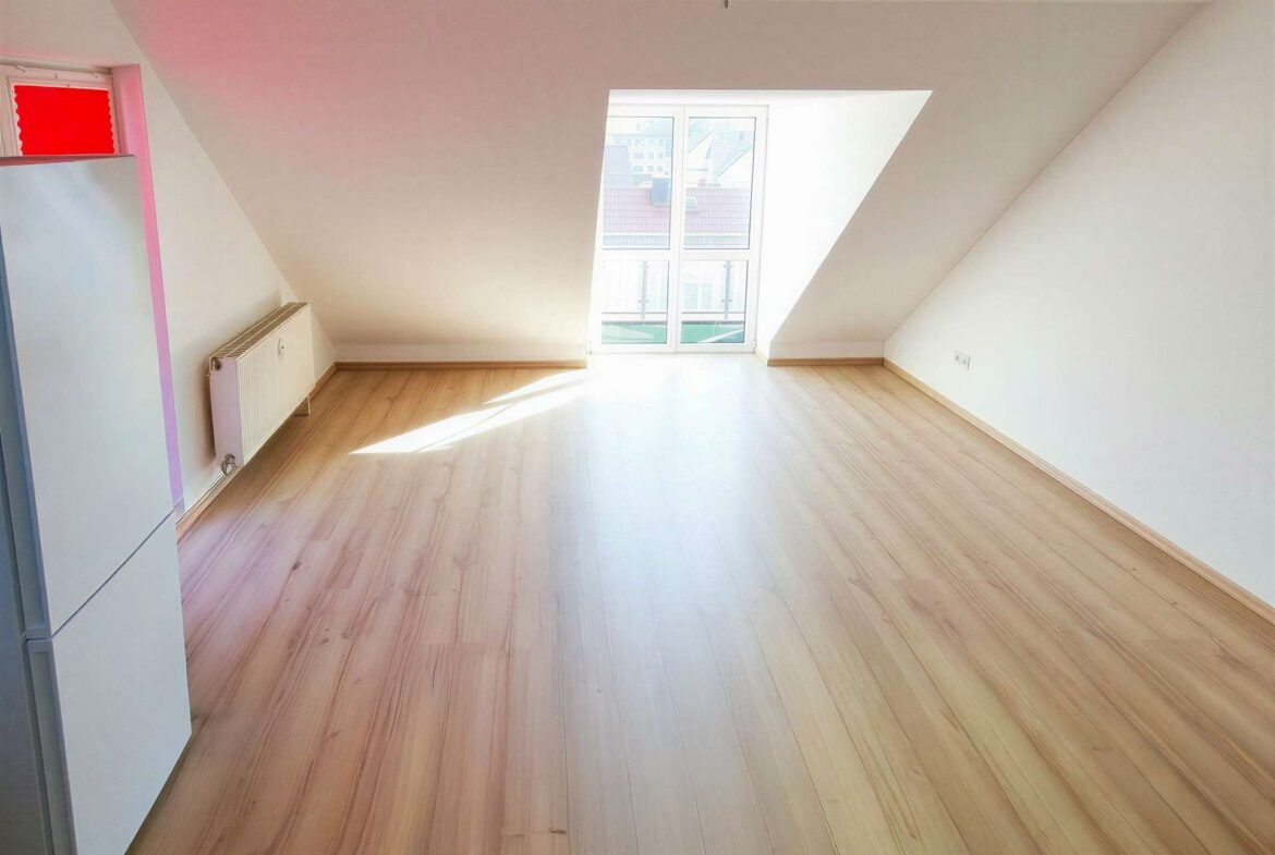 Bright empty attic room with sloped walls, a large window at the far end, and a radiator along the left wall.