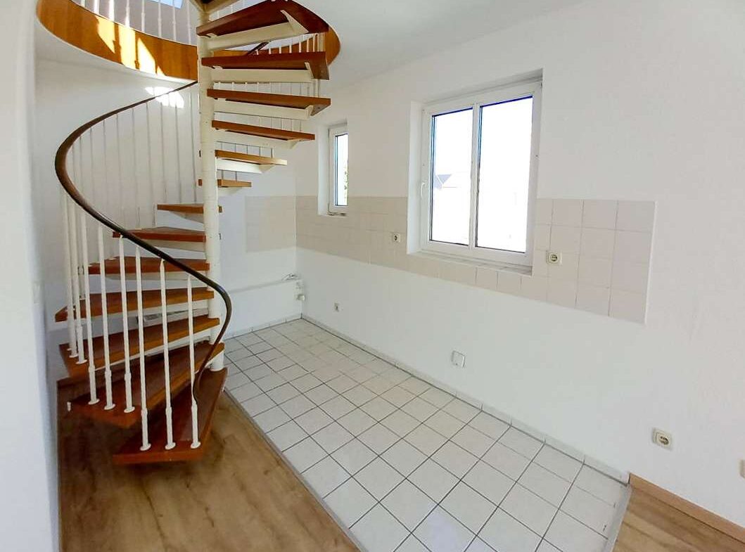 White-walled room with tiled floor and a wooden spiral staircase in the left corner, two windows along the back wall.