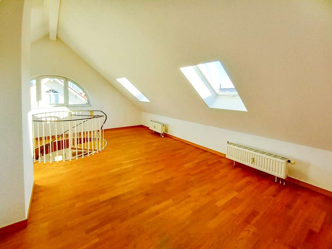 Bright attic room with sloped ceiling, two skylights, and a curved white railing by a spiral staircase; warm wooden floor and radiators along the wall.