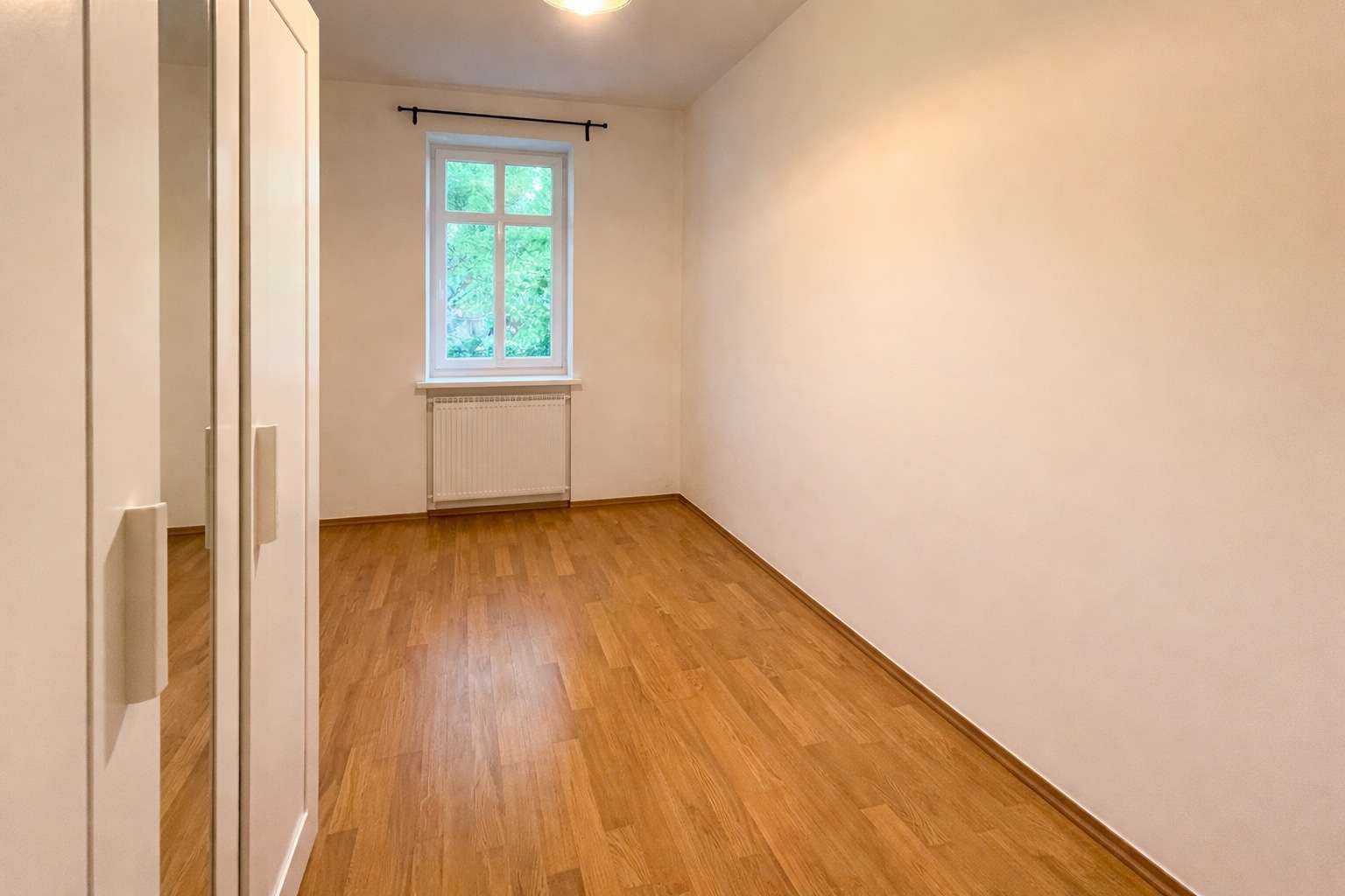 Narrow, empty bedroom with light wood floor, white wardrobe with mirrors on the left, window and radiator at the far wall.