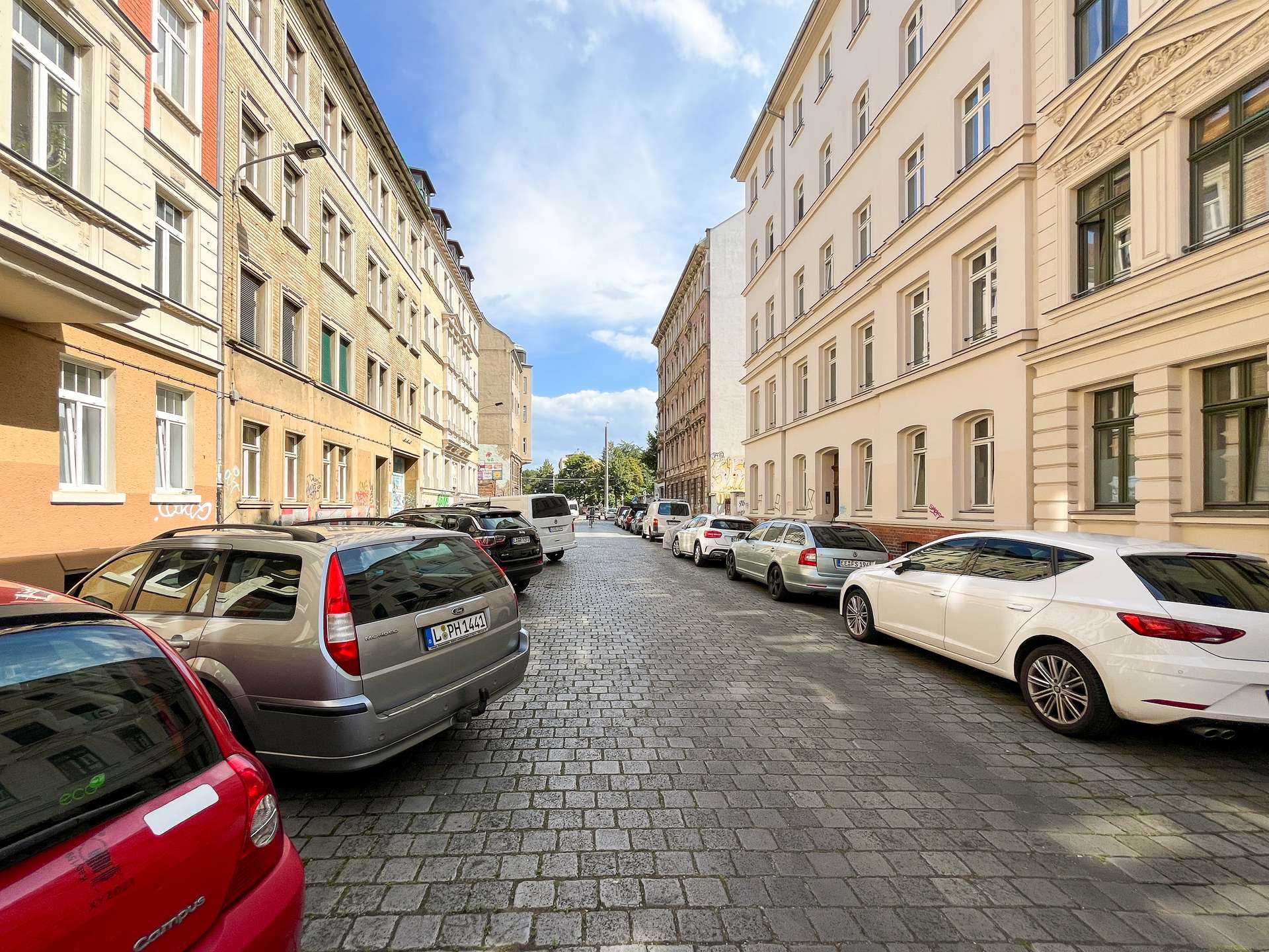 Narrow cobblestone street between light-colored apartment buildings, with cars parked on both sides under a blue sky.