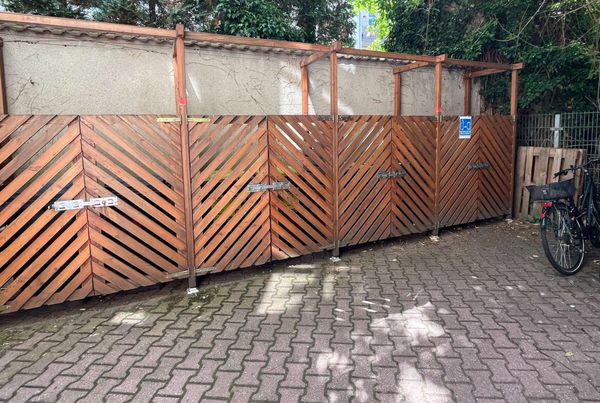 Row of wooden storage sheds with diagonal slat panels, metal latches, and a blue handicap sign; bicycle on the right.