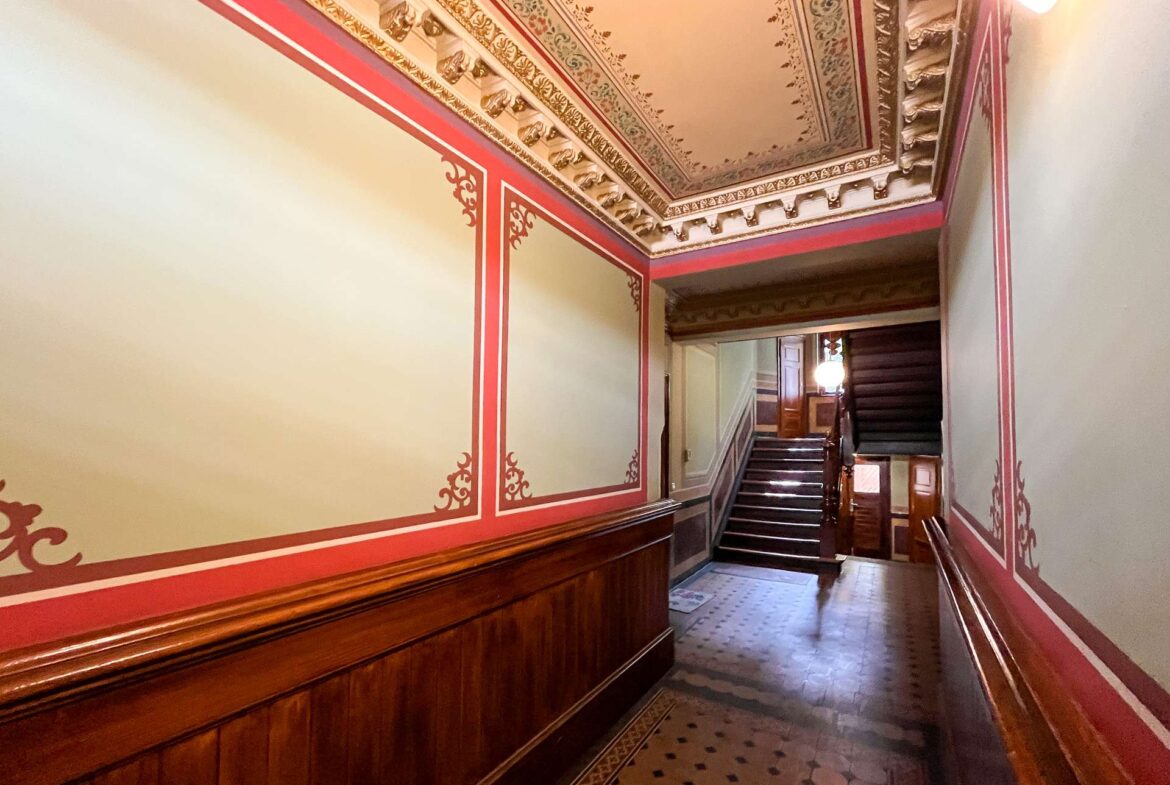 Grand historic hallway with ornate ceiling details, red wall trim, and dark wood wainscoting leading to a stairwell around the corner.