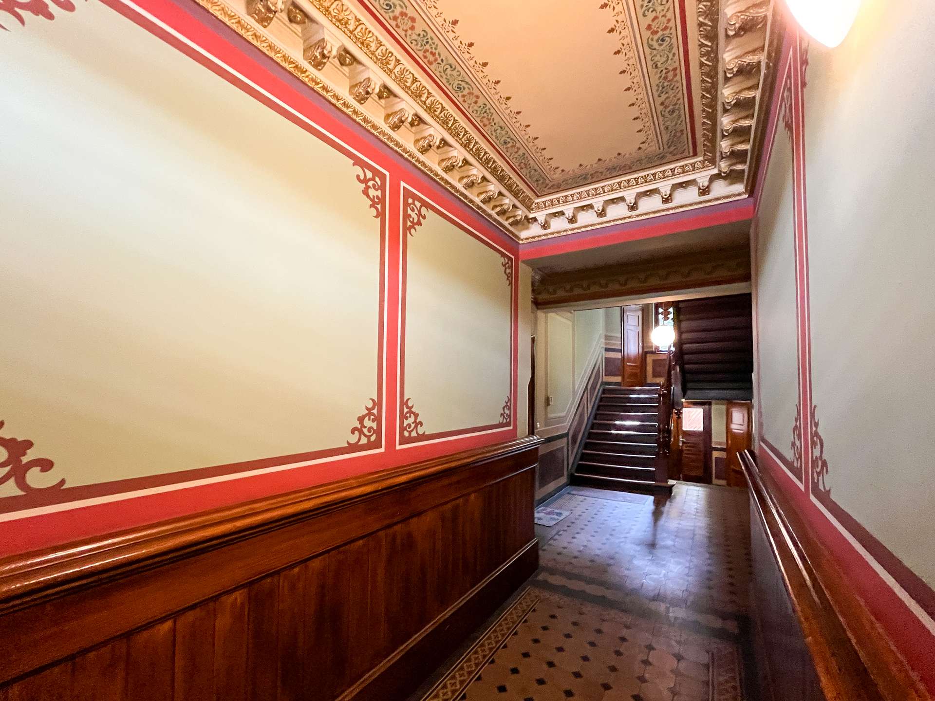 Grand historic hallway with ornate ceiling details, red wall trim, and dark wood wainscoting leading to a stairwell around the corner.