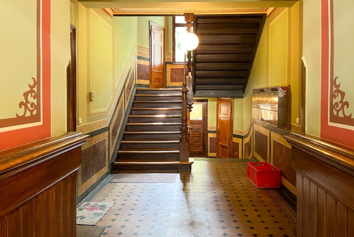 Interior staircase in a historic building: green walls with decorative trim, dark wooden banister, and a door at the landing.