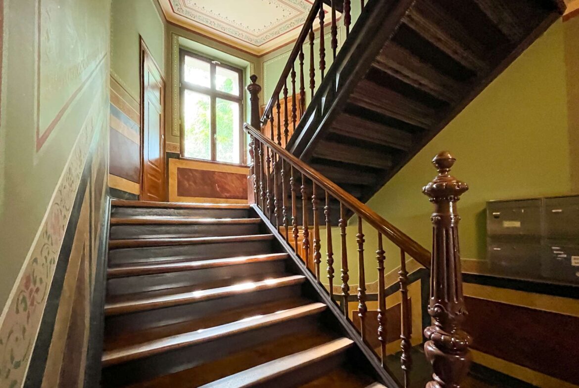 Wooden staircase with ornate balusters inside a green-walled stairwell, sunlight at the landing window.