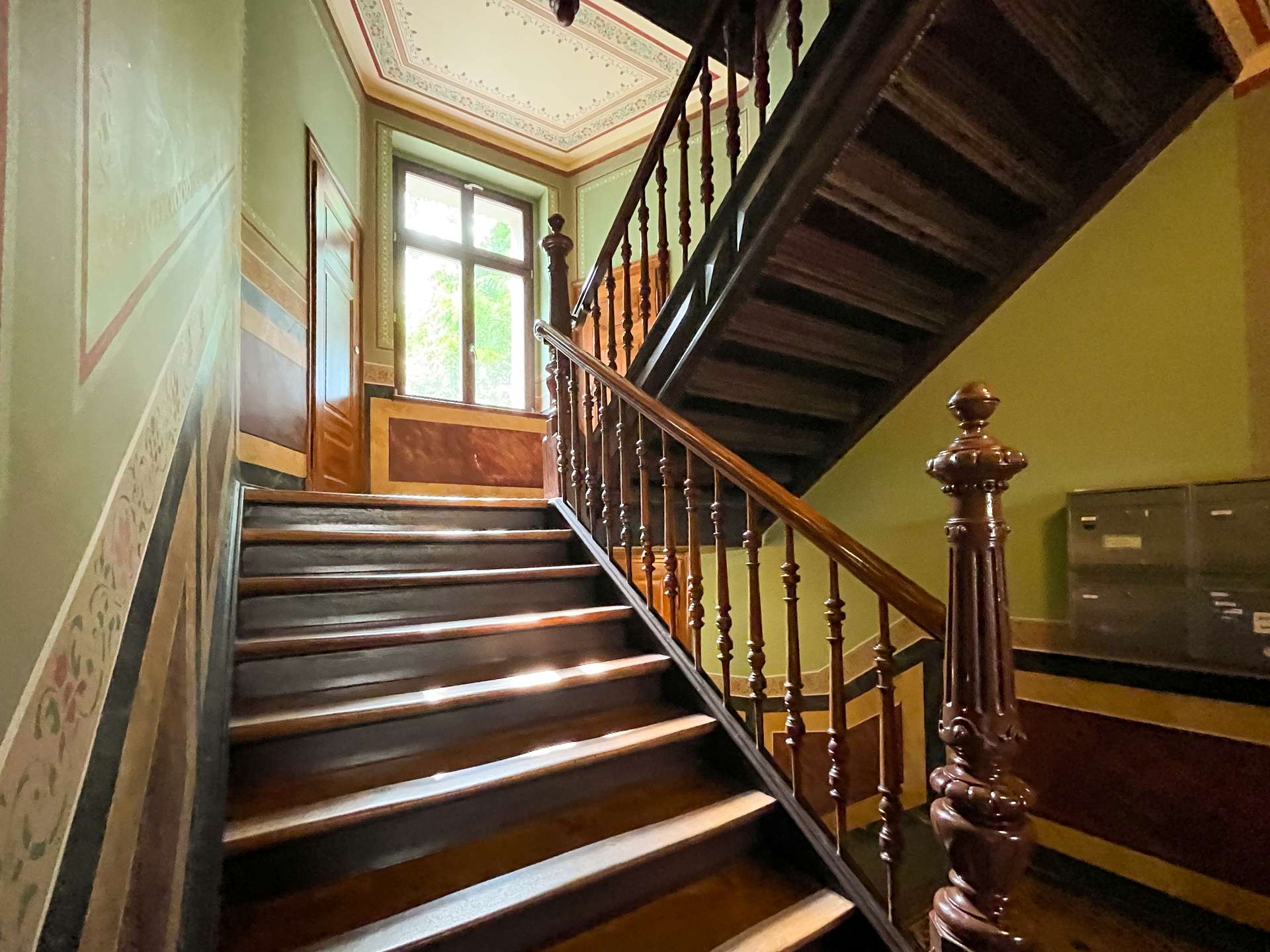 Wooden staircase with ornate balusters inside a green-walled stairwell, sunlight at the landing window.