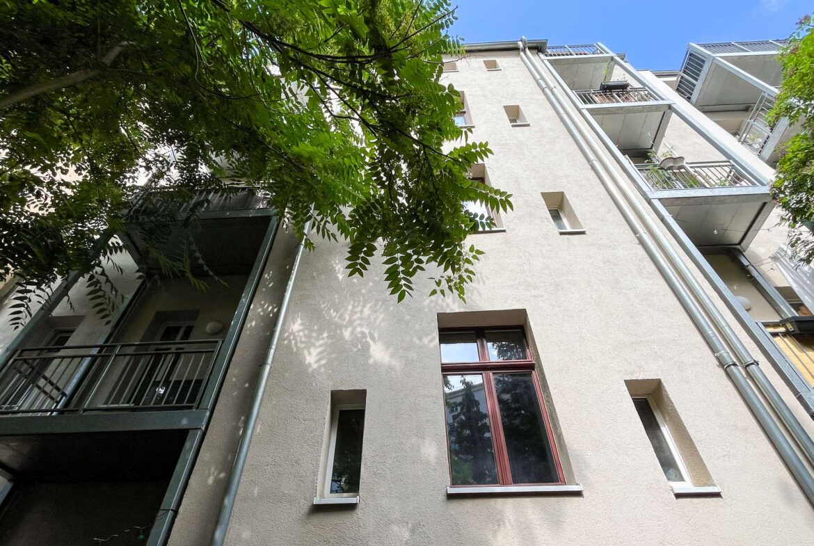 Low-angle view of a beige apartment building with balconies, metal railings, and vertical pipes, partially shaded by tree branches against a blue sky.