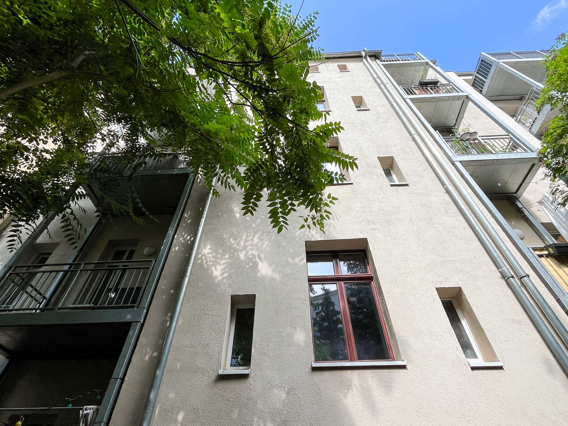 Low-angle view of a beige apartment building with balconies, metal railings, and vertical pipes, partially shaded by tree branches against a blue sky.