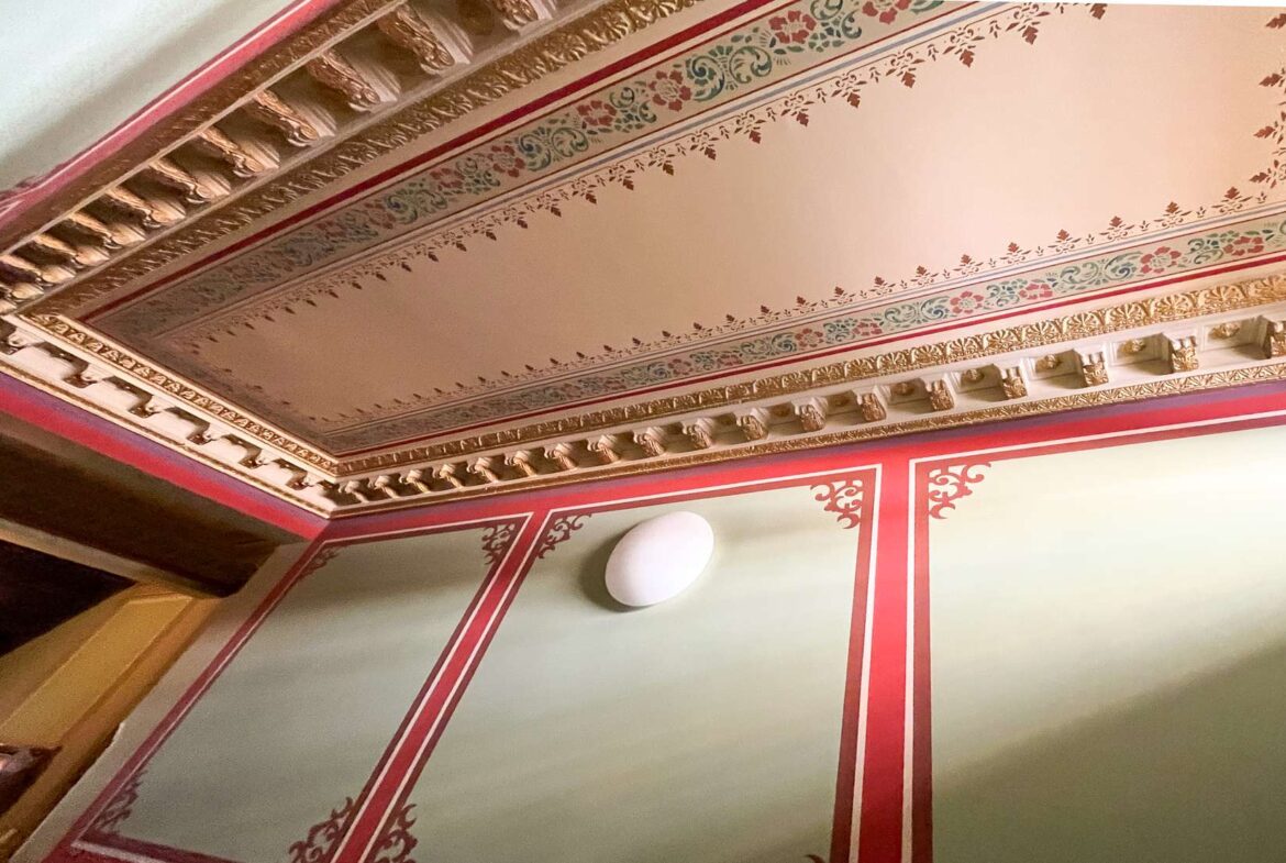 Ornate ceiling with gilded cornices, floral border patterns in red, gold, and green, and a white ceiling light fixture