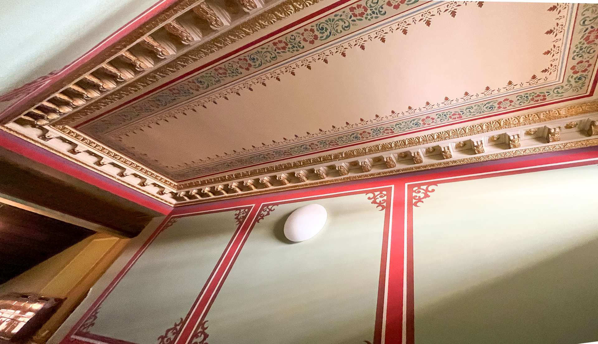 Ornate ceiling with gilded cornices, floral border patterns in red, gold, and green, and a white ceiling light fixture