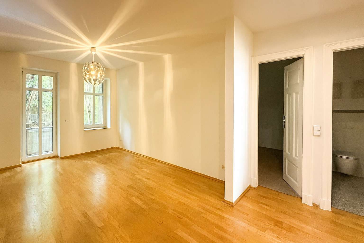 Bright empty living room with light wood floors, a decorative pendant light, and a glass door leading to an outdoor area.