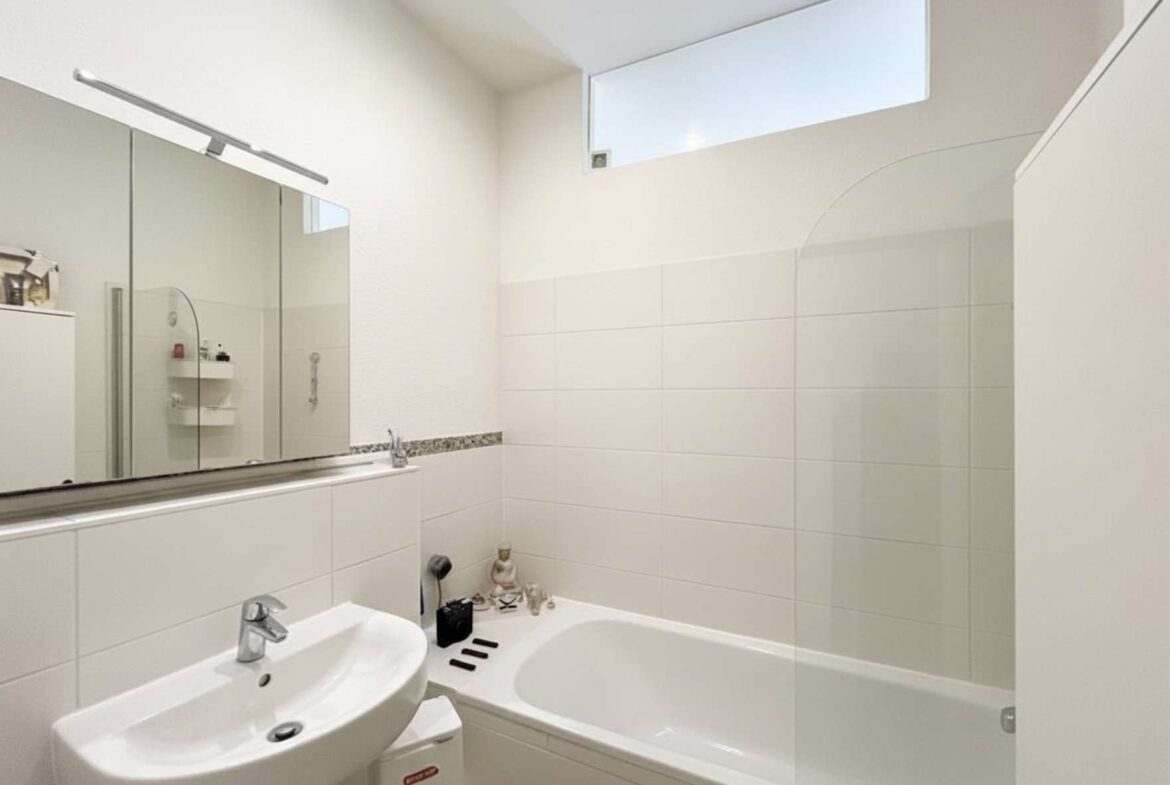 Modern white bathroom with a sink, pedestal cabinet and large mirror cabinet above; bathtub along the right with tiled walls and a small window above.