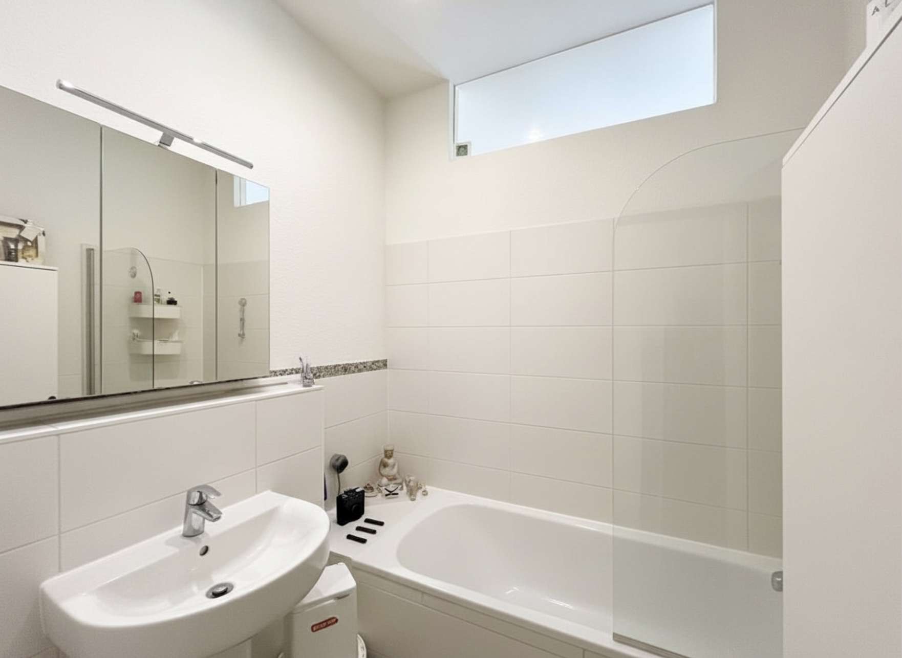 Modern white bathroom with a sink, pedestal cabinet and large mirror cabinet above; bathtub along the right with tiled walls and a small window above.