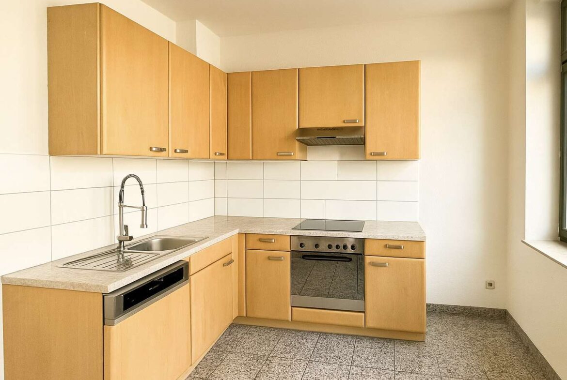 L-shaped kitchen with light wood cabinets, white tile backsplash, stainless steel sink, dishwasher, oven, and stovetop under a range hood. A window is on the right.