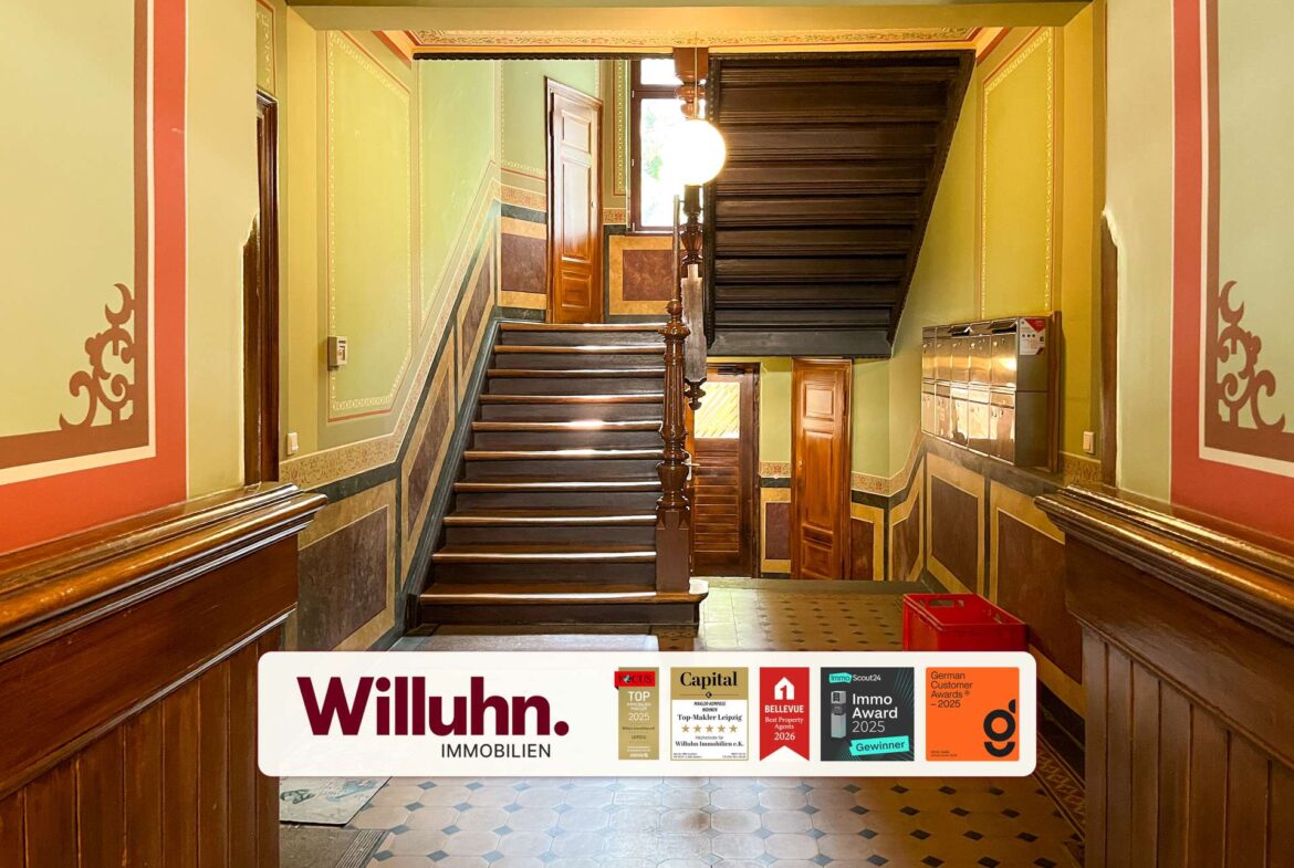 Historic building lobby with a dark wooden staircase, green walls with decorative trim, and a large Willuhn Immobilien banner at the bottom.